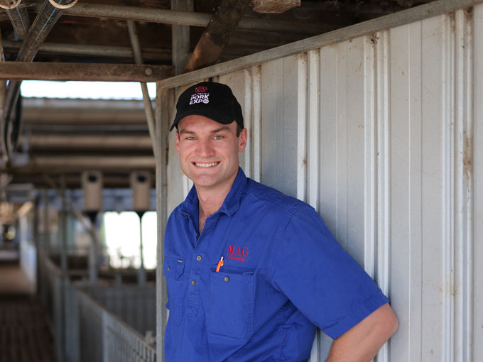 Farmer wearing blue shirt leaning against wall of pig shed. Smiling at camera.